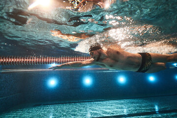 sports advertising underwater photography of a swimmer in a pool swimming freestyle in a sports cap, swimming goggles and dark swimming trunks - obrazy, fototapety, plakaty