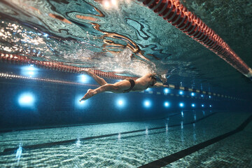 sports advertising underwater photography of a swimmer in a pool swimming freestyle in a sports cap, swimming goggles and dark swimming trunks - obrazy, fototapety, plakaty