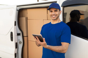 Delivery driver man smiling at camera beside his van full of parcel boxes, holding digital tablet,...