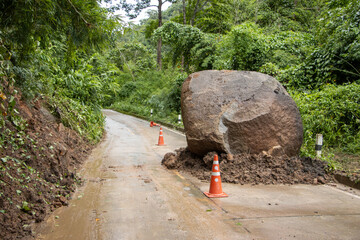 Large boulders fall from the mountain, blocking a rural concrete road in a green forest during the rainy season. There are orange cones placed on the road. Large rocks fall and soil slides