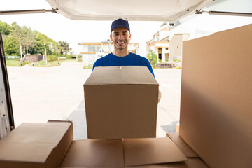 Young man courier taking out cardboard box packages from opened delivery van to give postal parcel to customer