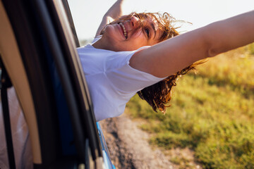 Young woman leans out of car window and laughs. Smiling girl travels and has fun on road