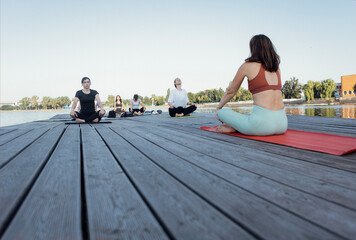Outdoor yoga class with an instructor near the river. Young women sit cross-legged on mats and...