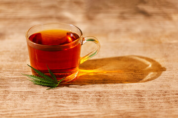 A transparent cup of delicious strong black tea with a cannabis leaf. A glass cup and hemp on a wooden table. A healthy plant-based organic drink.