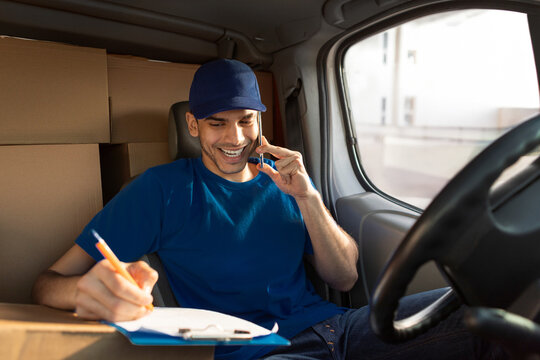 Cheerful european courier man calling clients with smartphone and checking address of delivery sitting in van full of cardboard boxes