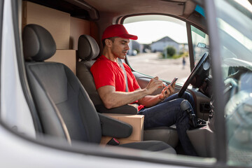 Young delivery man courier sitting in car and using mobile smartphone checking location to contact...