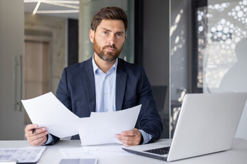 Portrait of a serious young man of a financial expert, the director of a company sitting in the office at the table in a suit, holding documents in his hands and confidently looking at the camera