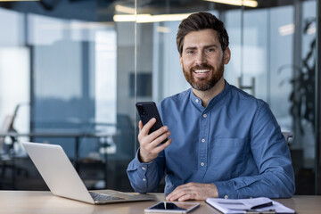 Portrait of a young man who works in the office at a laptop, holds a phone in his hand and looks at the camera with a smile