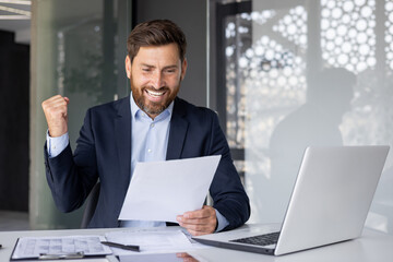 Happy male businessman looking at documents and enjoying success, loving victory gesture, sitting...