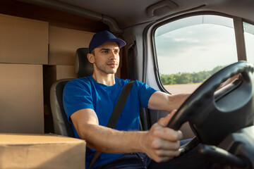 Young serious delivery man in uniform sitting inside van full of cardboard boxes, delivering...