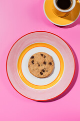 Chocolate chip cookie on a bright plate, pink backdrop with a cup of coffee.