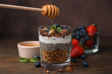 Pouring honey onto tasty granola with berries, yogurt and chia seeds at wooden table