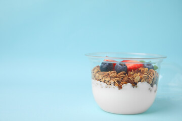 Tasty granola with berries and yogurt in plastic cup on light blue background, space for text