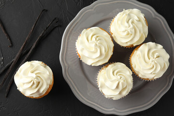 Tasty cupcakes with cream and vanilla pods on black table, top view