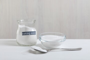 Baking soda in bowl, glass jar and spoon on white wooden table