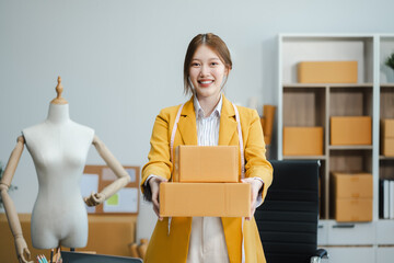 Young Asian SMEs woman holding parcels and looking to camera, standing among several boxes in...