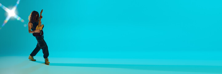 Obraz premium Young guy with log hair playing electric guitar, expressing energy against blue, cyan background in neon. Concept of music, performance, festival, live concert. Banner. Empty space to insert text