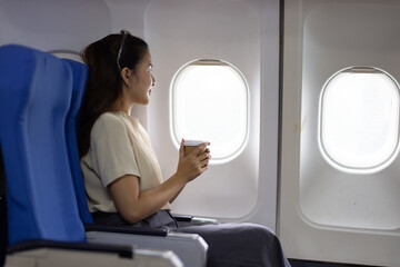 Young Asian female tourist traveling alone on a plane is drinking hot coffee and looking out the window.