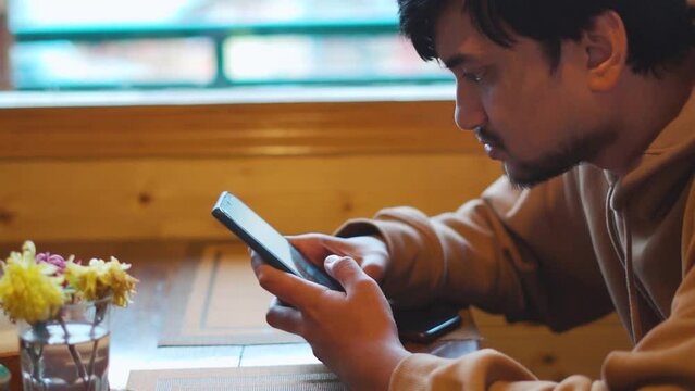 Closeup shot of an Indian man scrolling his mobile phone while sitting inside cafe and waiting for food. Man addicted to mobile phone. Man scrolls the phone while waiting. Social media addiction. 