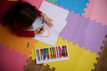 Little girl smiling drawing picture, lying on her belly on a multicolored puzzle carpet. People....