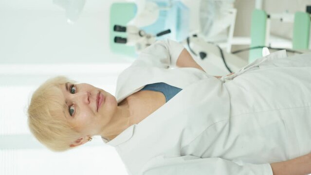 Vertical video portrait of a female gynecologist doctor in a white coat. An examination chair and a colposcope can be seen in the background. High quality 4k footage