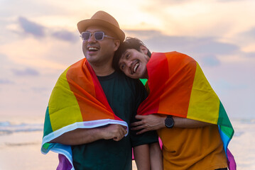 Happy Gay couple with lgbt flag looking to sunset together on the beach in summer day. Happy guys demonstrate their rights. LGBTQI, Pride Event, LGBT Pride Month, Gay Pride Symbol.