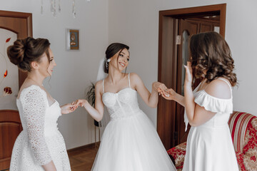 Three women are standing in a room, one of them is wearing a wedding dress. They are all smiling...