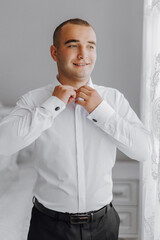 A man in a white shirt is getting dressed and smiling. He is wearing a black belt and black pants
