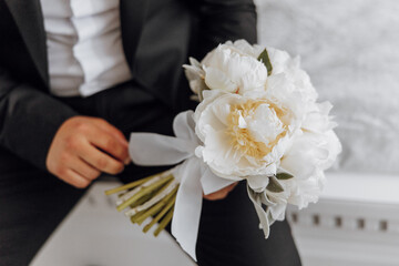 A man is sitting on a chair holding a bouquet of white flowers. The man is wearing a suit and tie,...