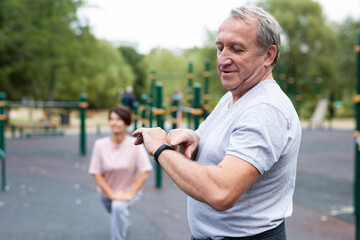 Ssenior man in sportswear looks at fitness watch or at pedometer while exercising in city park - 800249521