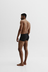 Fototapeta premium Young African-American man with relief strong back, muscular body standing shirtless in black boxers isolated on white background. Concept of male beauty, sport, body care, health, fitness