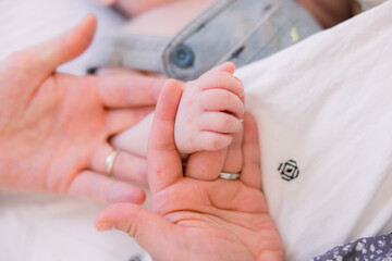 A married father is holding the hand of his newborn baby boy, who is wearing overalls. 
