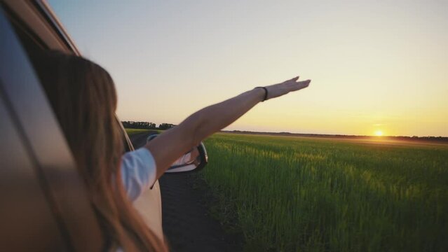 Woman catches wind with hand from car window at sunset in countryside. Blonde girl with long hair stretching arm out catching glare of setting sun. Romantic car trip, adventure tourism concept.