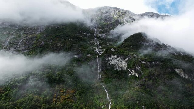 Incredible thin waterfall descends cascading between clouds and forest in Milford Sound, aerial parallax