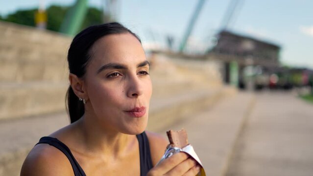 Athletic female pausing for a snack, indulging in a chocolate protein bar to refuel post-exercise. Energetic jogger taking a moment for nutrition against the backdrop of a cityscape