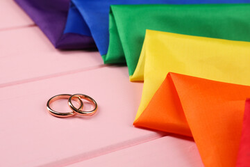 Rainbow LGBT flag and wedding rings on pink wooden table, closeup