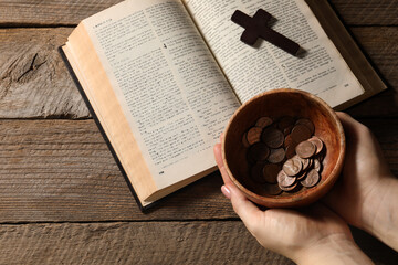 Donate and give concept. Woman holding bowl of coins. Bible and cross on wooden table, top view