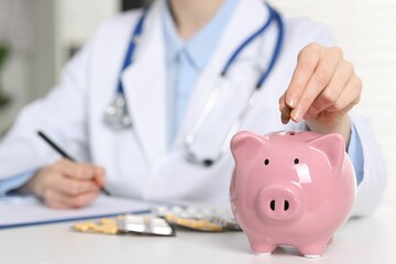 Doctor putting coin into piggy bank at white table indoors, closeup