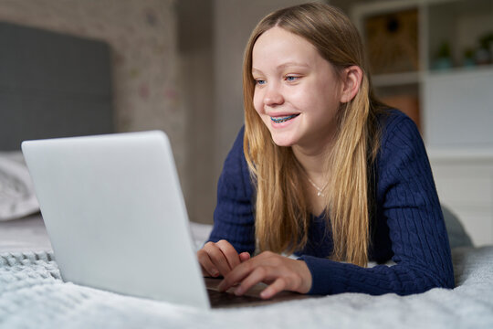 Teenage Girl Wearing Orthodontic Braces With Laptop Lying On Bed At Home Gaming, Streaming Film Or Show, Browsing Online Looking At Social Media