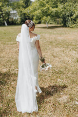 A bride is walking in a field with a white veil and flowers in her hands. The scene is serene and peaceful, with the bride looking towards the camera