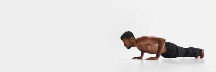 Young handsome African-American man training in pants shirtless, doing push ups isolated on white background. Male beauty, sport, body care, health, fitness concept. Banner. Empty space to insert text
