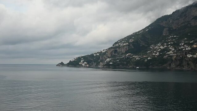 Smooth panning video along Italy&rsquo;s Amalfi Coast, featuring steep mountains rising from the Mediterranean Sea, coastal villages, dramatic cliffs, and homes built into the rugged hillside beneath a clou