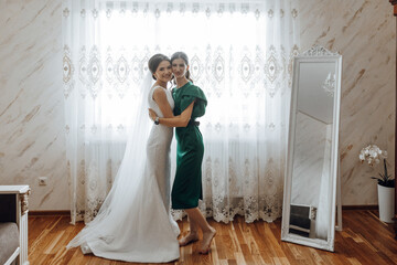 A bride and her mother are standing in front of a mirror. The bride is wearing a white dress and...