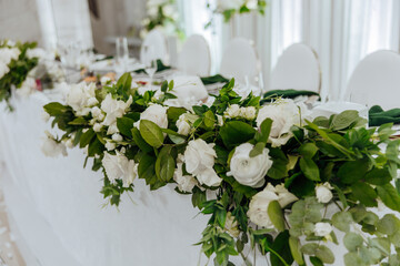 A table with white flowers and green leaves. The table is set for a special occasion. The flowers are arranged in a way that they look like they are cascading down the table
