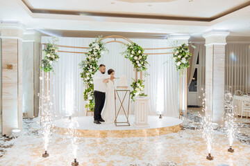 A bride and groom are cutting a cake in front of a white arch. The arch is decorated with flowers...