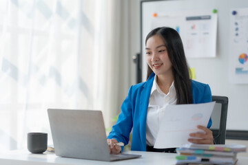Beautiful young Asian business woman manager or company worker holding accounting document, checking financial data or marketing report working in office with laptop.