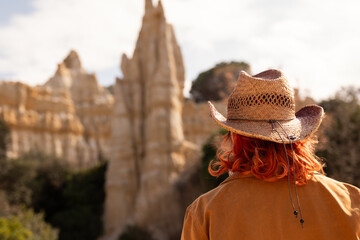 A woman wearing a brown jacket and a hat stands in front of a large rock formation. Concept of adventure and exploration, as the woman is enjoying her time in the desert