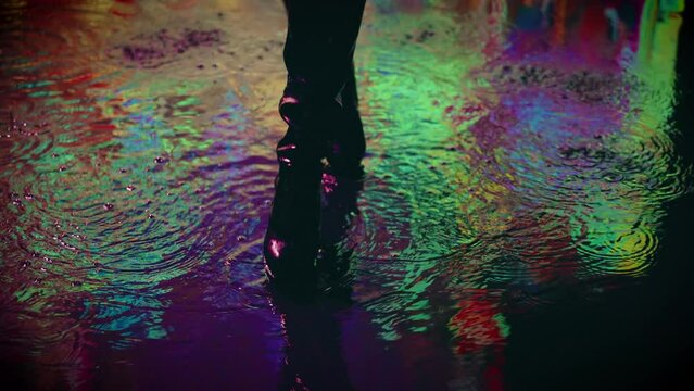 unrecognizable female legs feet close-up stepping through puddles wet road adult girl walks in rain night city neon light reflected in water. woman walking in black boots fashion model in aqua studio