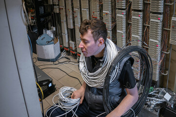 A technician with a bundle of cables around his neck works in a server room. A man lays a cable at a telephone exchange - obrazy, fototapety, plakaty