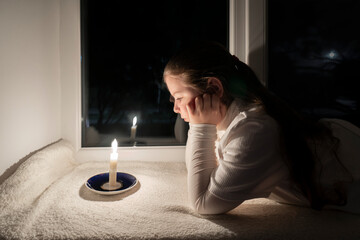 Blackout. A girl in a dark room lies on the windowsill near the window and looks at a burning candle